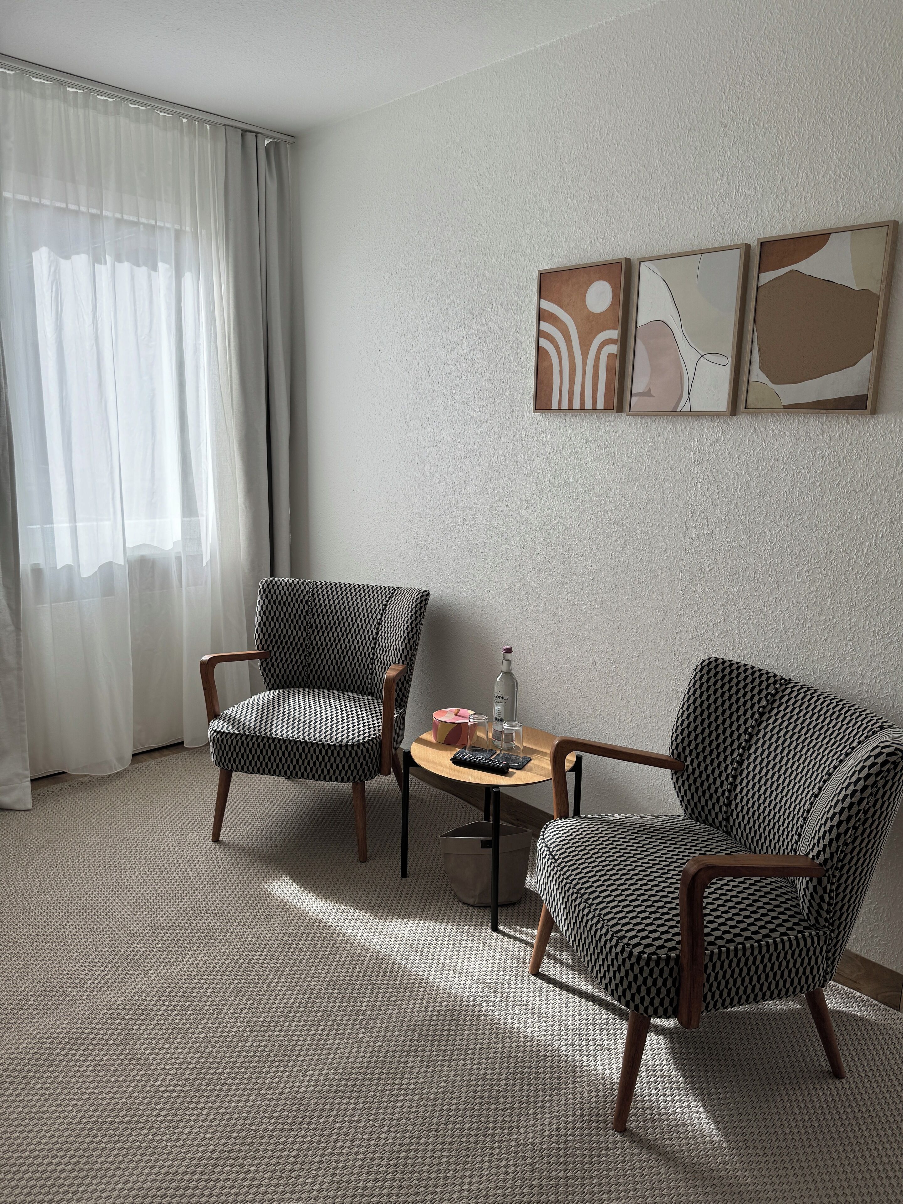 A room with two patterned armchairs, a small table with a bottle and a glass, and a window with sheer curtains.