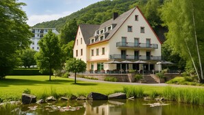 A large, multi-story hotel with a prominent balcony area, situated next to a calm pond with lily pads. - Hotel Alte Mühle (Bad Bertrich)