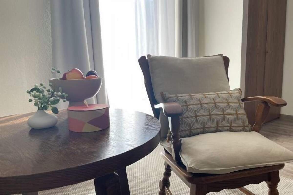 A round wooden table with a bowl of fruit and a vase of flowers, a patterned armchair with a cushion, and a window with curtains.