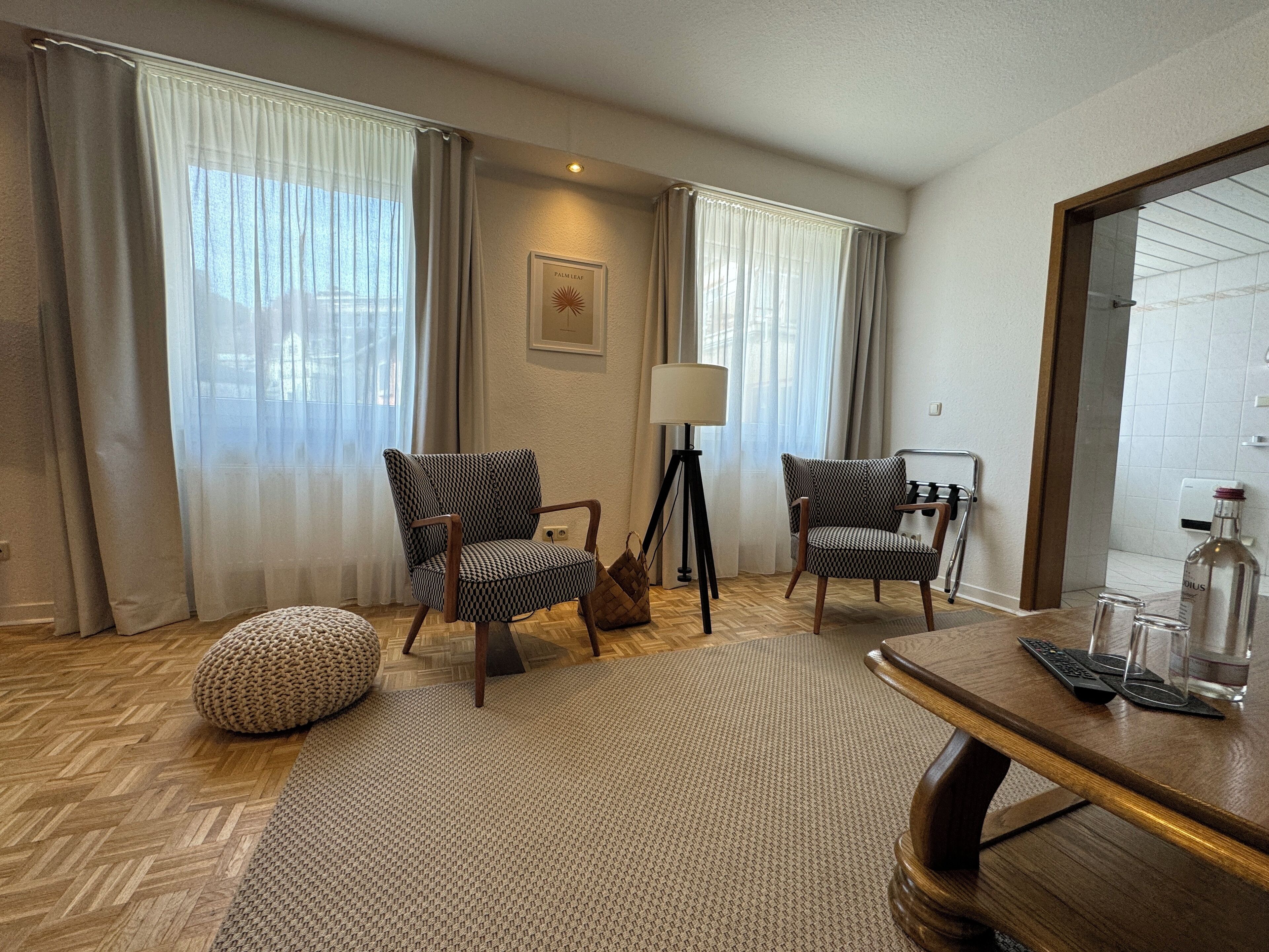 Una habitación de hotel con dos sillones estampados, una lámpara de pie, una mesa de centro de madera y un espejo grande.