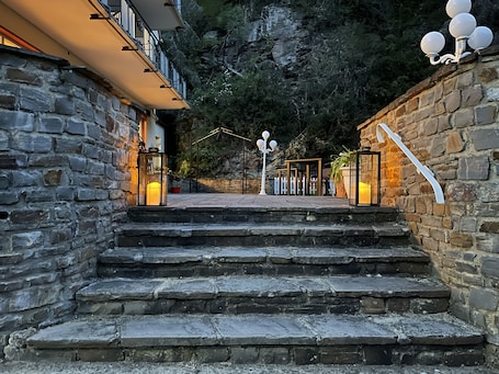A stone staircase leading to a building with a balcony, surrounded by a stone wall and a view of a rocky hillside.