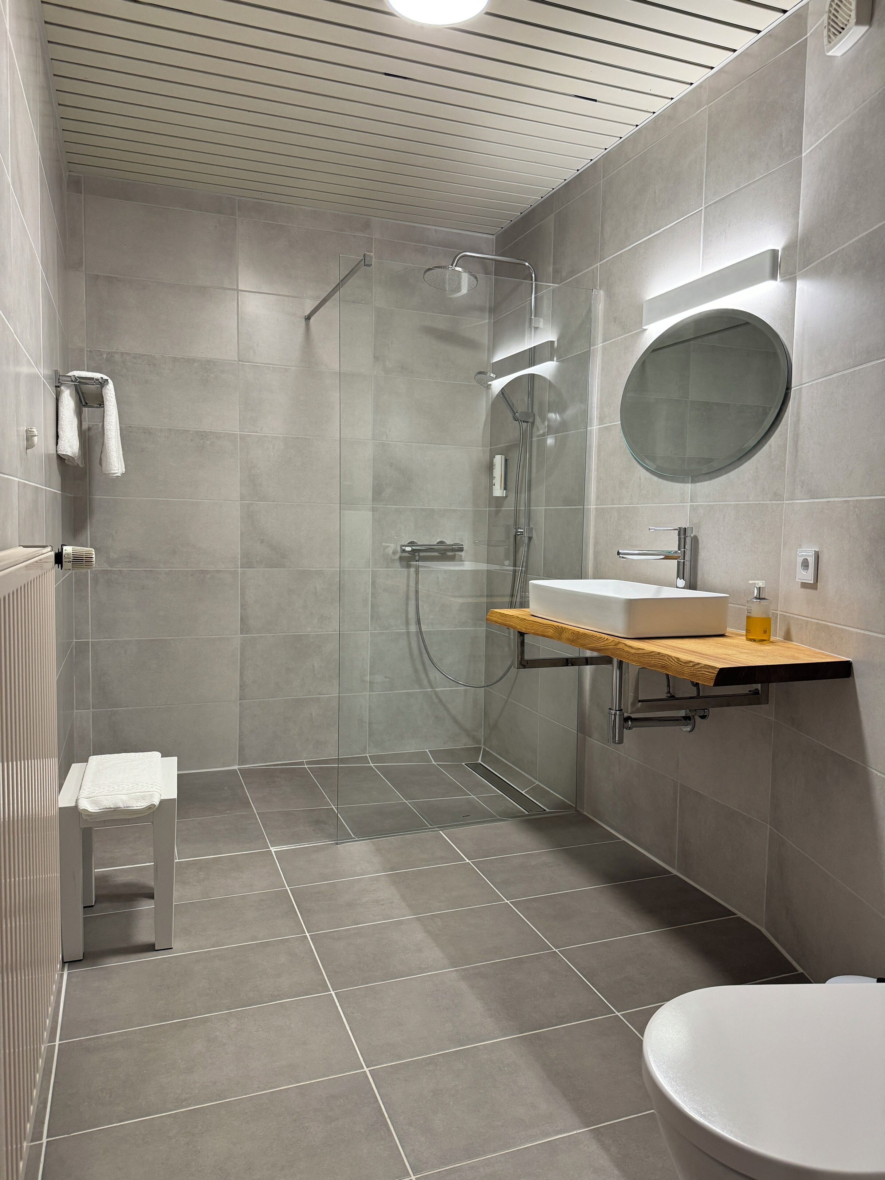 A modern bathroom with a glass-enclosed shower, a wooden vanity, a round mirror, and a toilet.