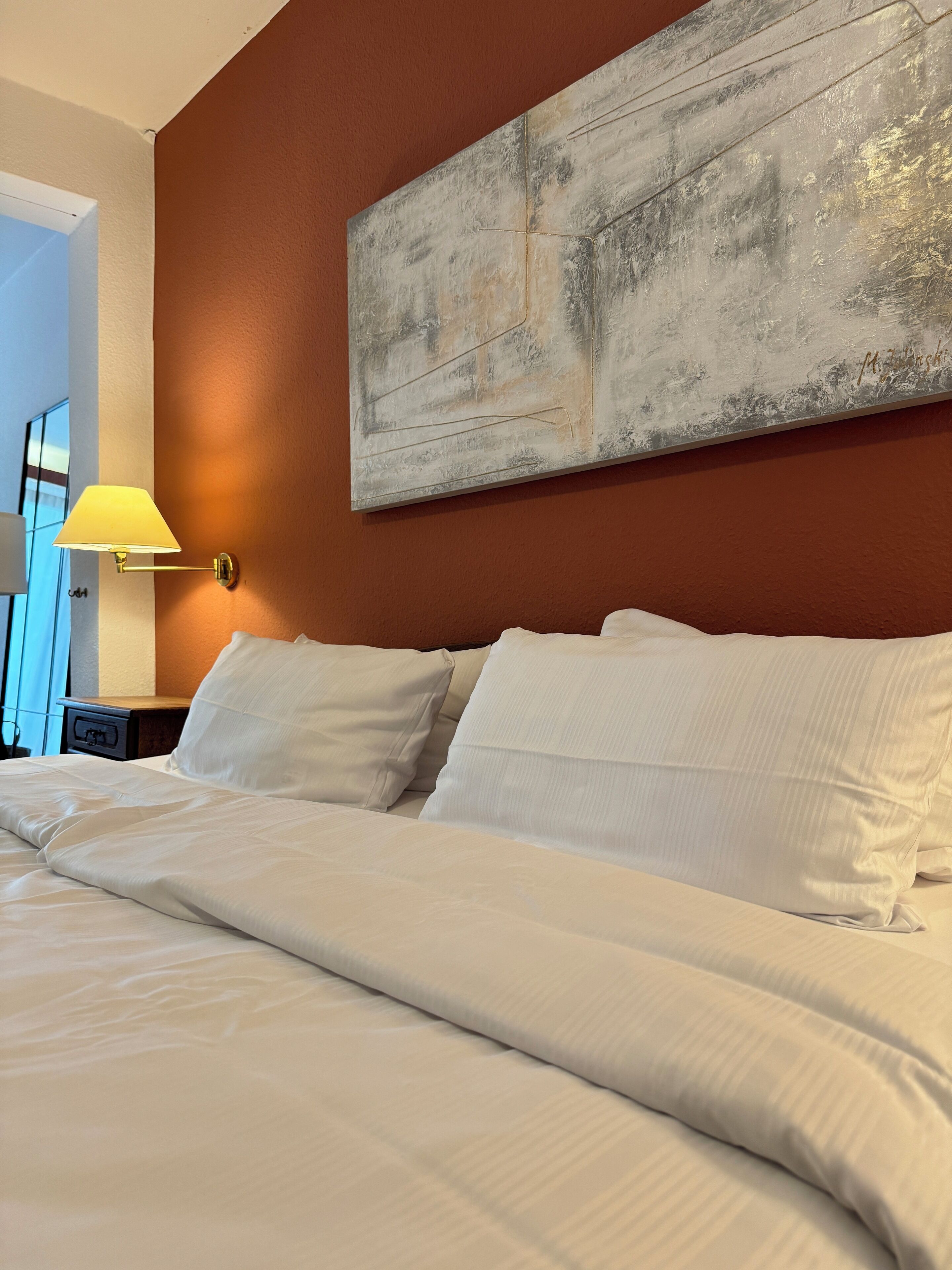 Una habitación de hotel con una cama grande, ropa de cama blanca, un cuadro abstracto colgado en la pared y una mesita de noche con una lámpara.