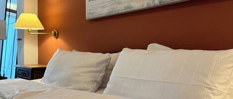 Una habitación de hotel con una cama grande, ropa de cama blanca, un cuadro abstracto colgado en la pared y una mesita de noche con una lámpara.