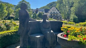 A garden with a stone sculpture of two figures, a building in the background, and a clear blue sky. - Hotel Alte Mühle (Bad Bertrich)