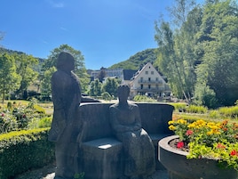 Un jardín con una escultura de piedra de dos figuras, un edificio al fondo y un cielo azul despejado.