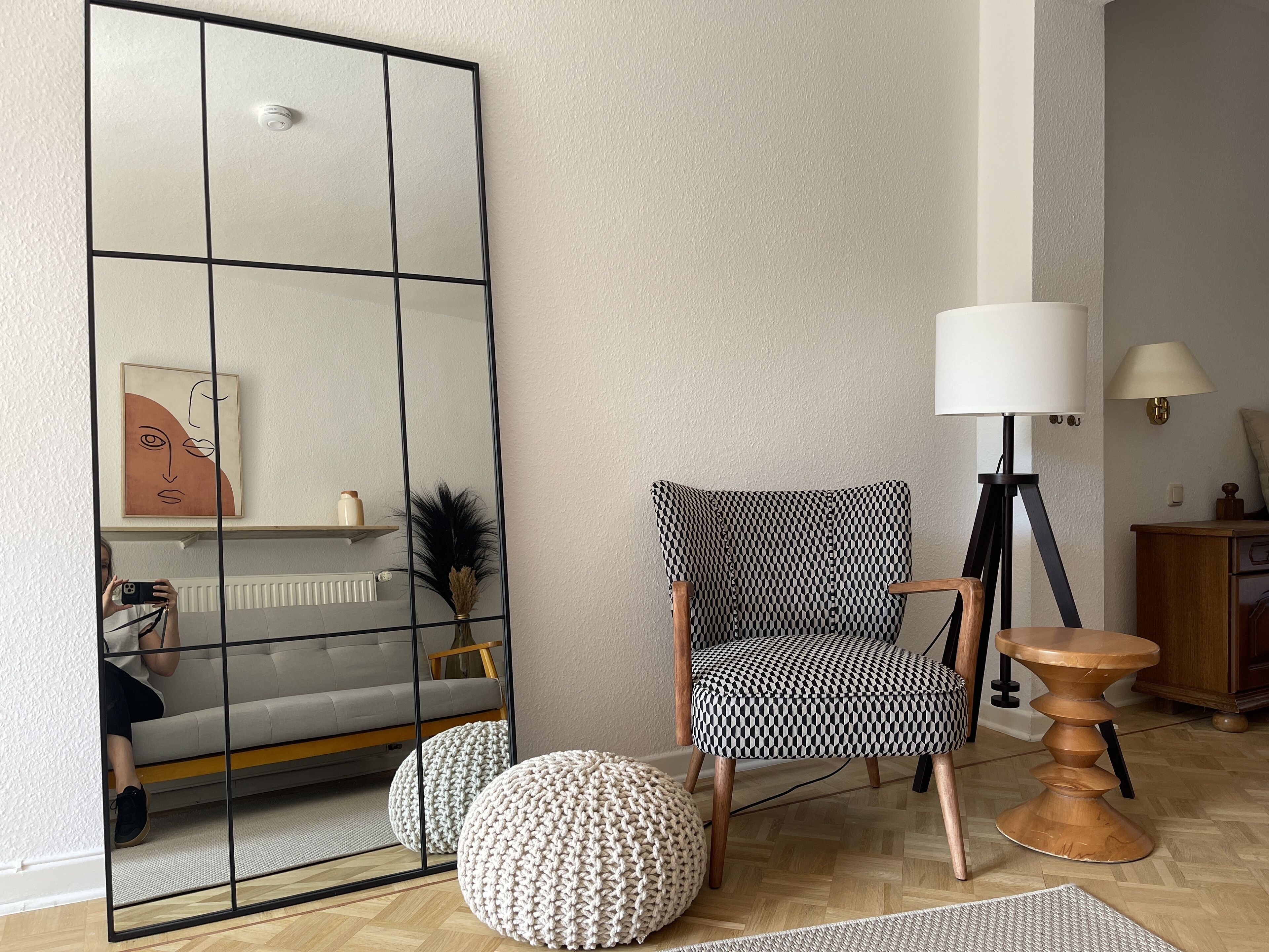 A modern living room with a large mirror, a patterned armchair, a round pouf, a wooden side table, and a floor lamp.