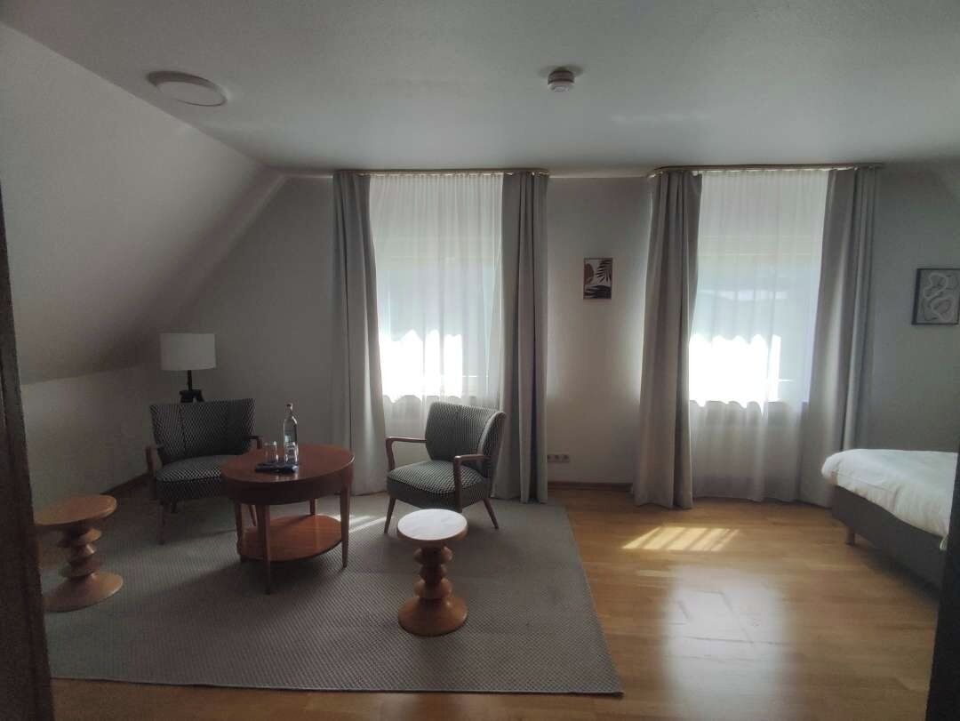 A hotel room with a bed, a round table, two chairs, a lamp, and a view of the outdoors through large windows.