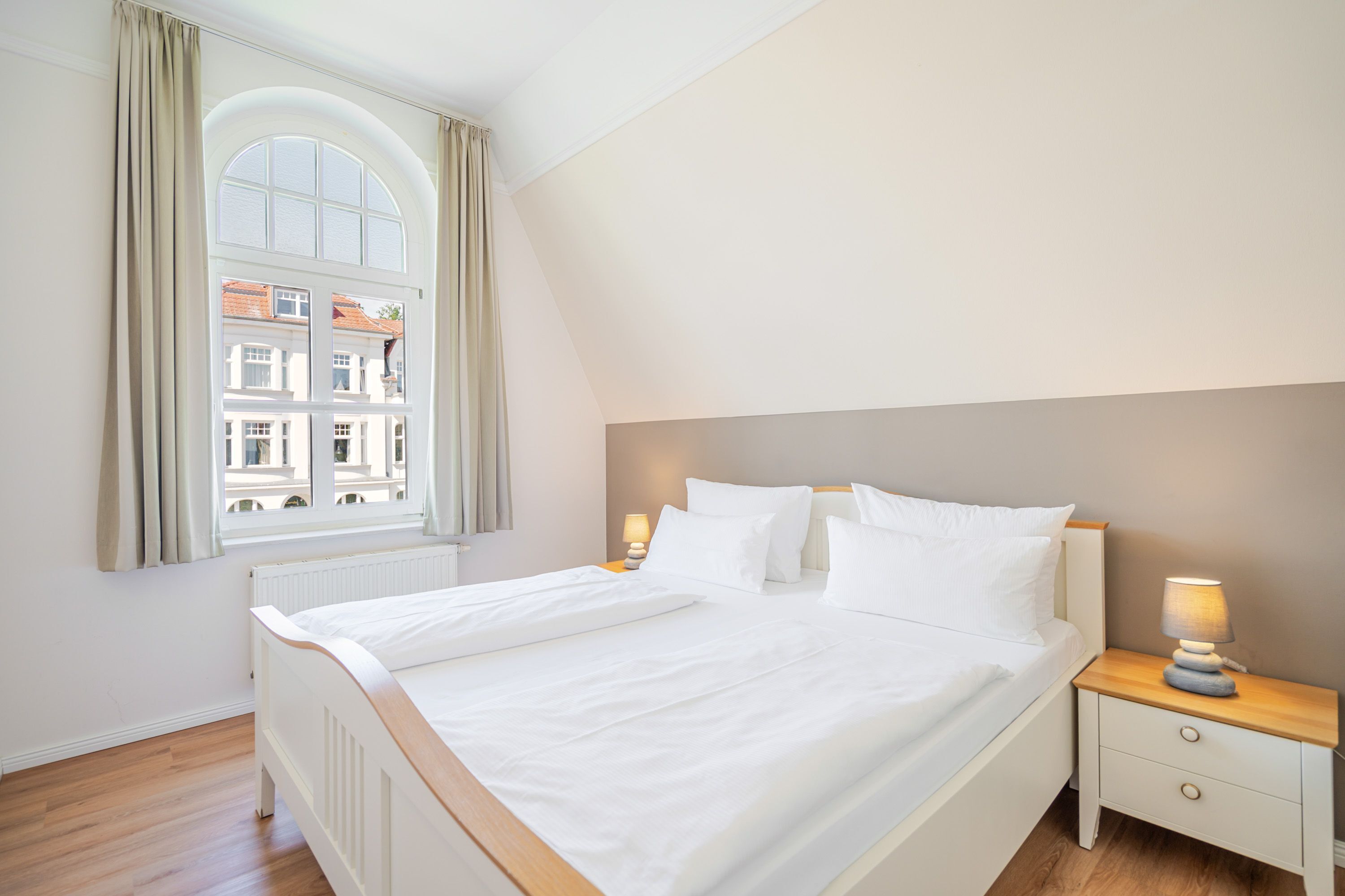 Un dormitorio con un amplio ventanal de arco, una cama con ropa de cama blanca, una mesita de noche con lámpara y suelo de madera.