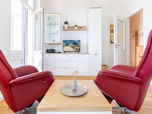 Ein modernes Wohnzimmer mit einem Holztisch, zwei roten Stühlen, einem Fernseher auf einem weißen Schrank und einem Glastisch mit Dekorationselementen.