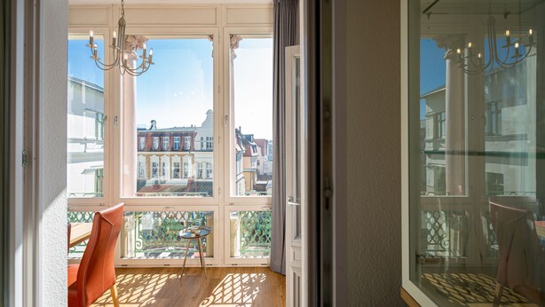 Una stanza con pavimento in legno, una sedia rossa e vista sugli edifici attraverso ampie finestre.