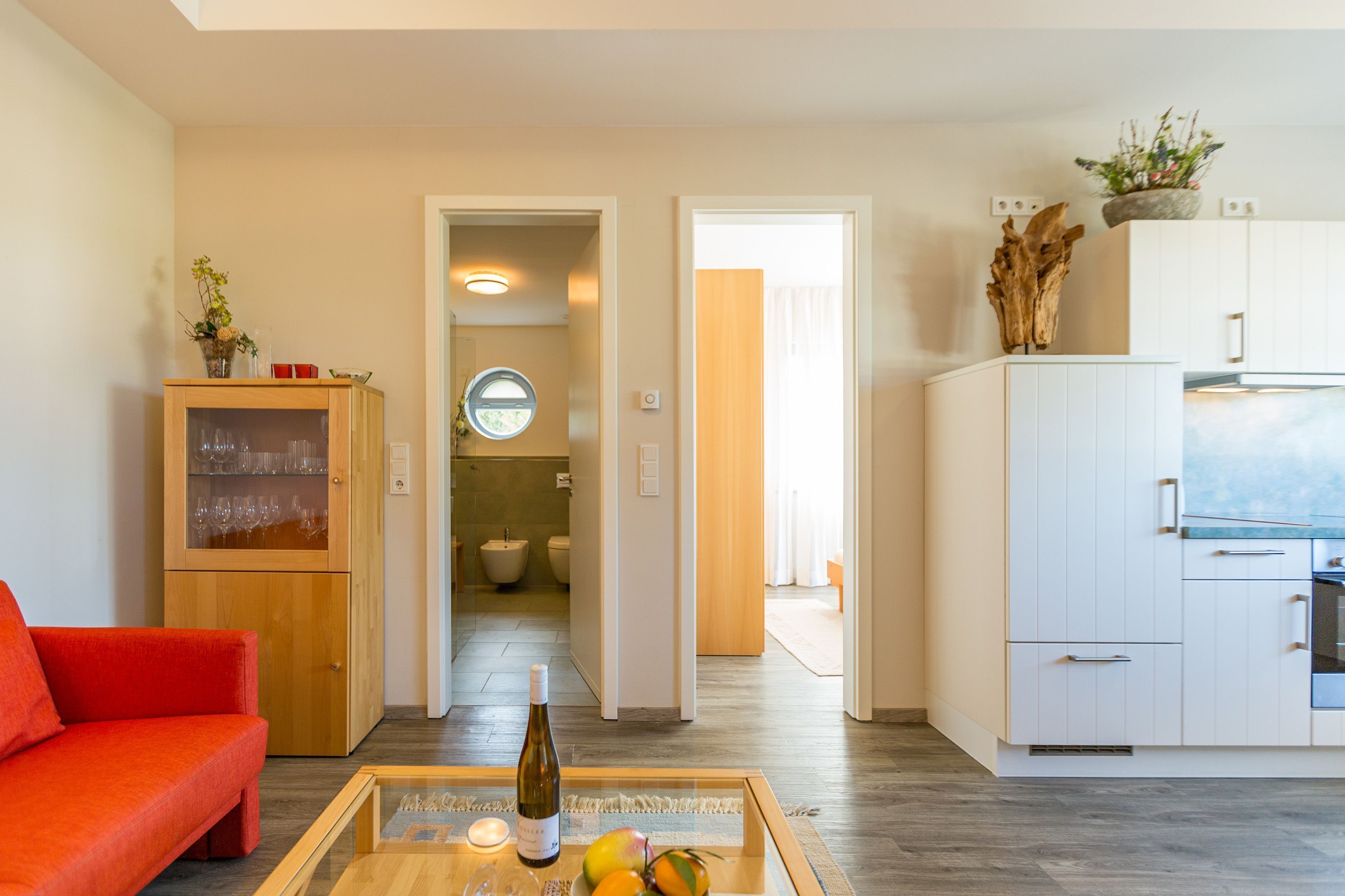 Een moderne woonkamer met een rode bank, een houten kast met glazen deuren, een keuken met witte kasten, en een gang naar een badkamer.