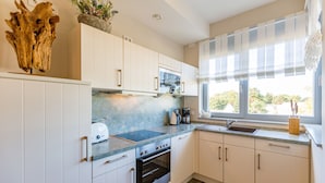 A modern kitchen with white cabinets, a built-in oven, a microwave, and a window with a view of greenery. - Quartier II, App. 2.2 (Heringsdorf)
