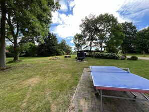 Een pingpongtafel buiten, in een gazon met bomen en een overdekte zithoek op de achtergrond.