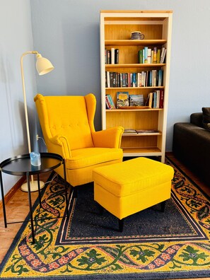 A room with a yellow armchair, a matching ottoman, a bookshelf, and a floor lamp.