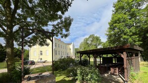 Une construction en briques surmontée d’une pergola en bois, une zone herbeuse et un bâtiment en arrière-plan.