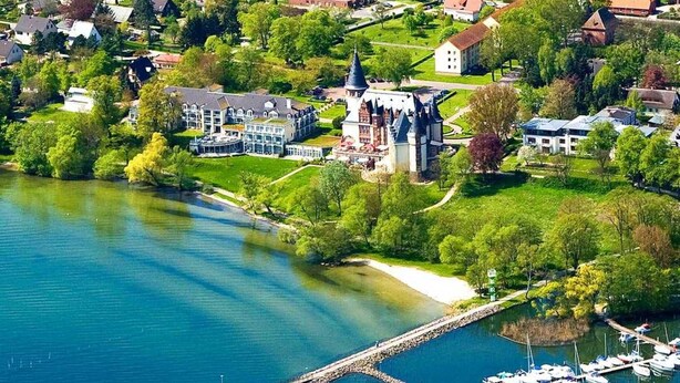 Vista aérea de un pueblo a orillas de un lago, con un edificio imponente de estilo castillo, un muelle con barcos y casas residenciales.