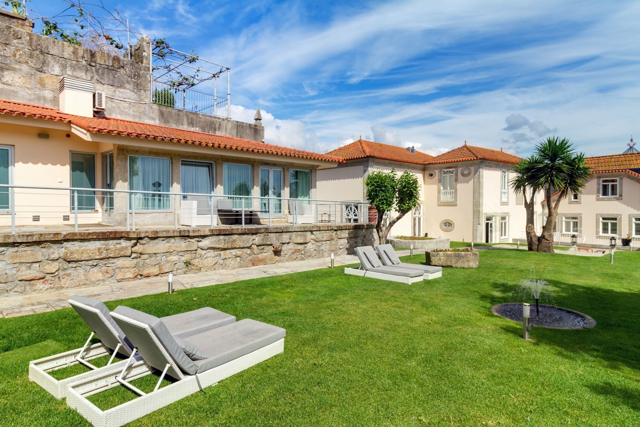A courtyard with lounge chairs, a fountain, and a building with a tiled roof and stone walls.