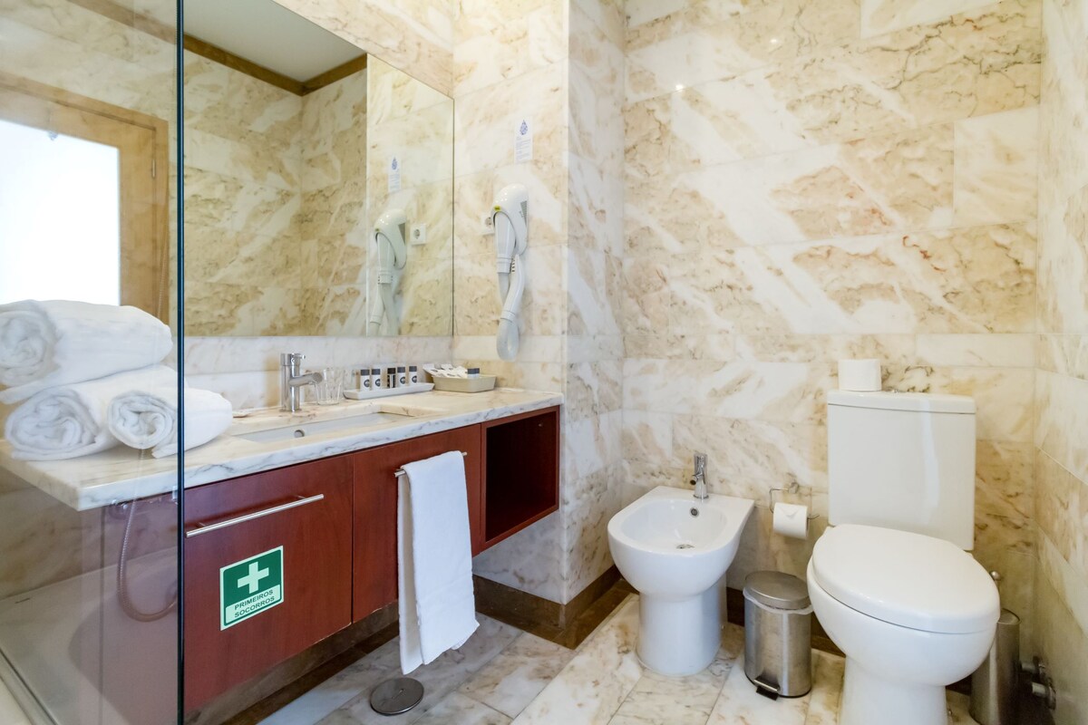 A bathroom with a marble wall, a glass-enclosed vanity, a bidet, a toilet, and a trash bin.