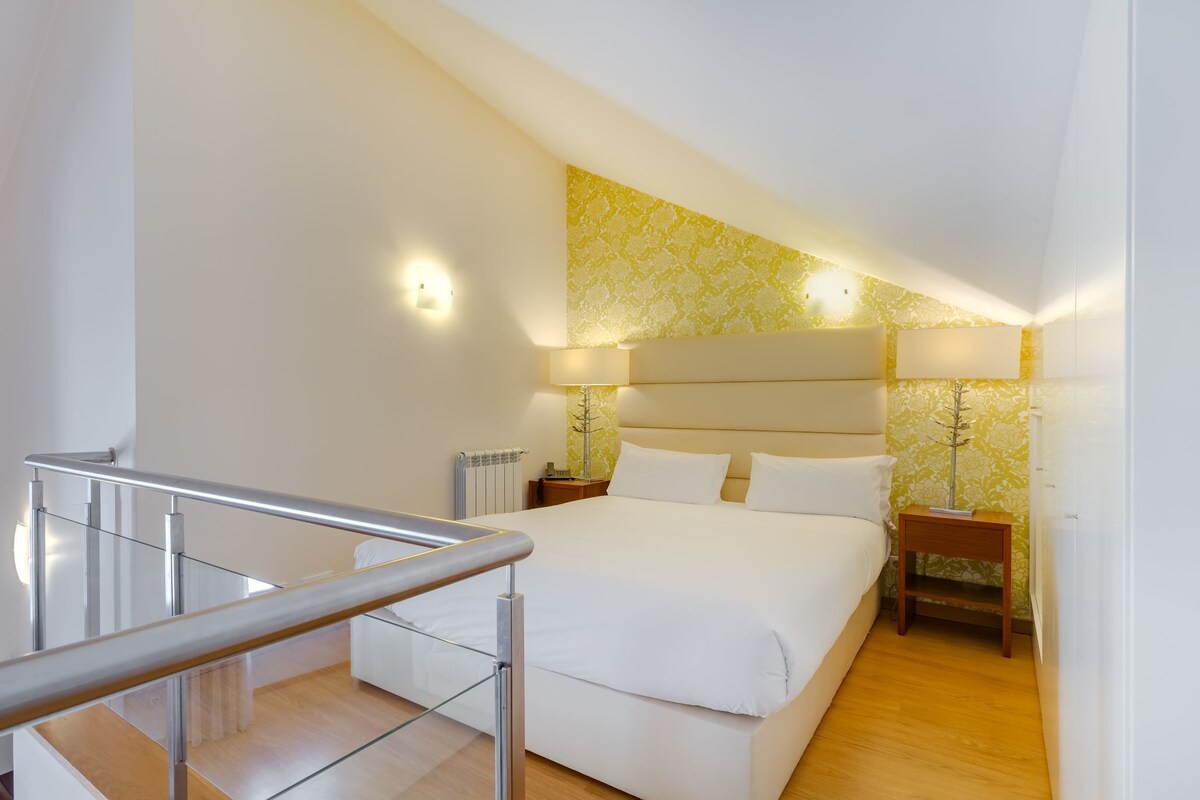 A modern hotel room with a large bed, a wooden floor, a glass railing, and a decorative wall.