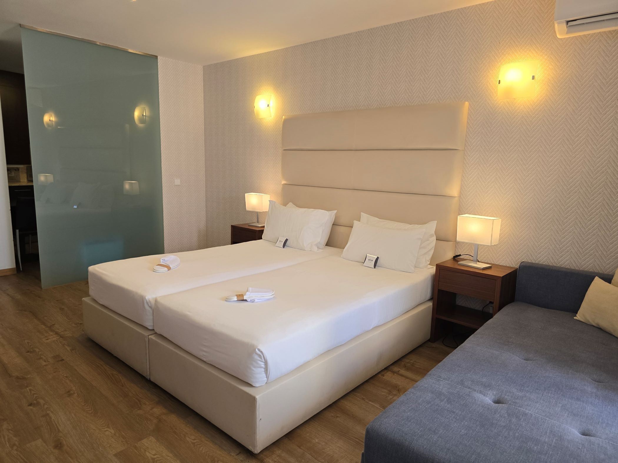 A hotel room with a large bed, two bedside tables with lamps, a grey sofa, and a herringbone-patterned wall.