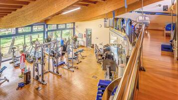 Ein Fitnessraum mit Holzbalken, Trainingsgeräten und großen Fenstern mit Blick auf eine Grünanlage.
