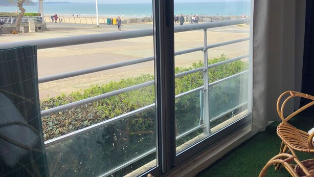 Quarto com uma grande janela com vista para a praia. Há uma cadeira de vime com almofada e uma pequena mesa.