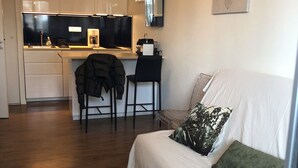 Uma sala de estar moderna com um sofá branco, uma mesa lateral redonda e uma área de cozinha ao fundo.