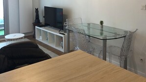 Uma sala de estar moderna com uma mesa de jantar de vidro, cadeiras transparentes, uma TV de tela plana e um lustre.