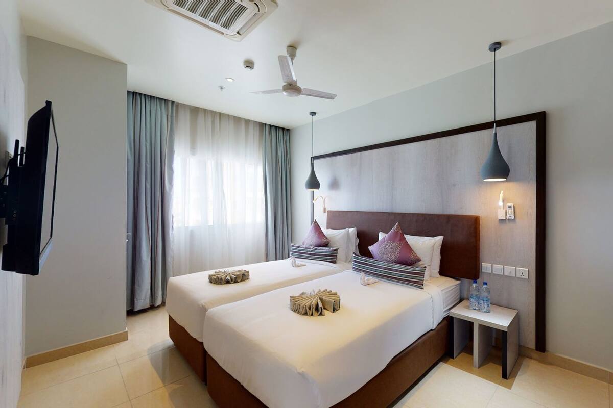 A hotel room with two beds, a ceiling fan, a TV, and a bedside table with a bottle.
