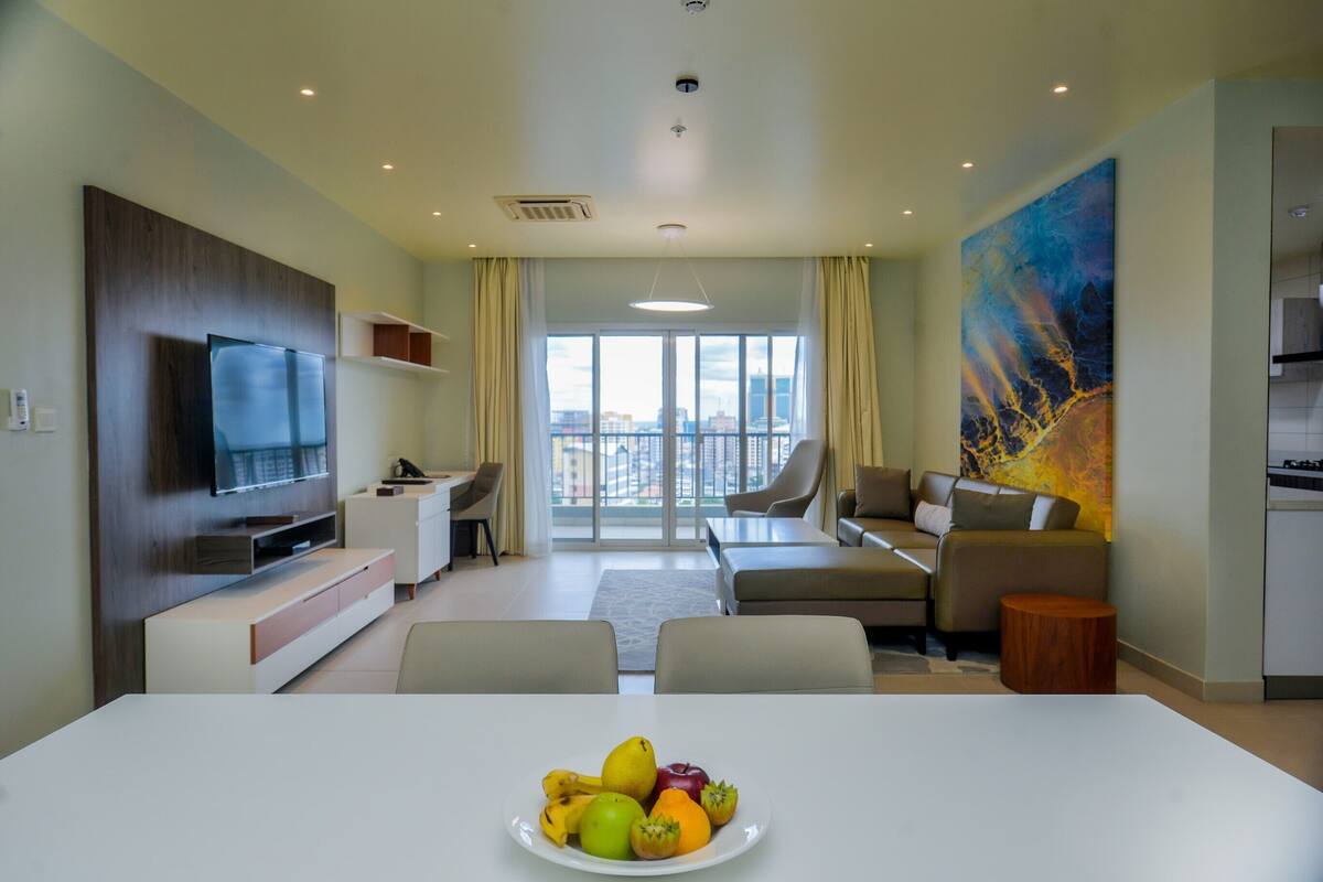 A modern living room with a flat-screen TV, a dining table with fruit, a sofa, and a large window with a city view.