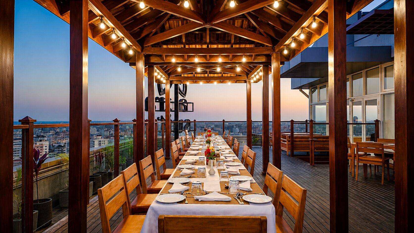 Un espacio de comedor al aire libre con una mesa larga dispuesta para una comida, vigas de madera y vista a la ciudad.