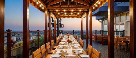 An outdoor dining area with a long table set for a meal, wooden beams, and a city view.