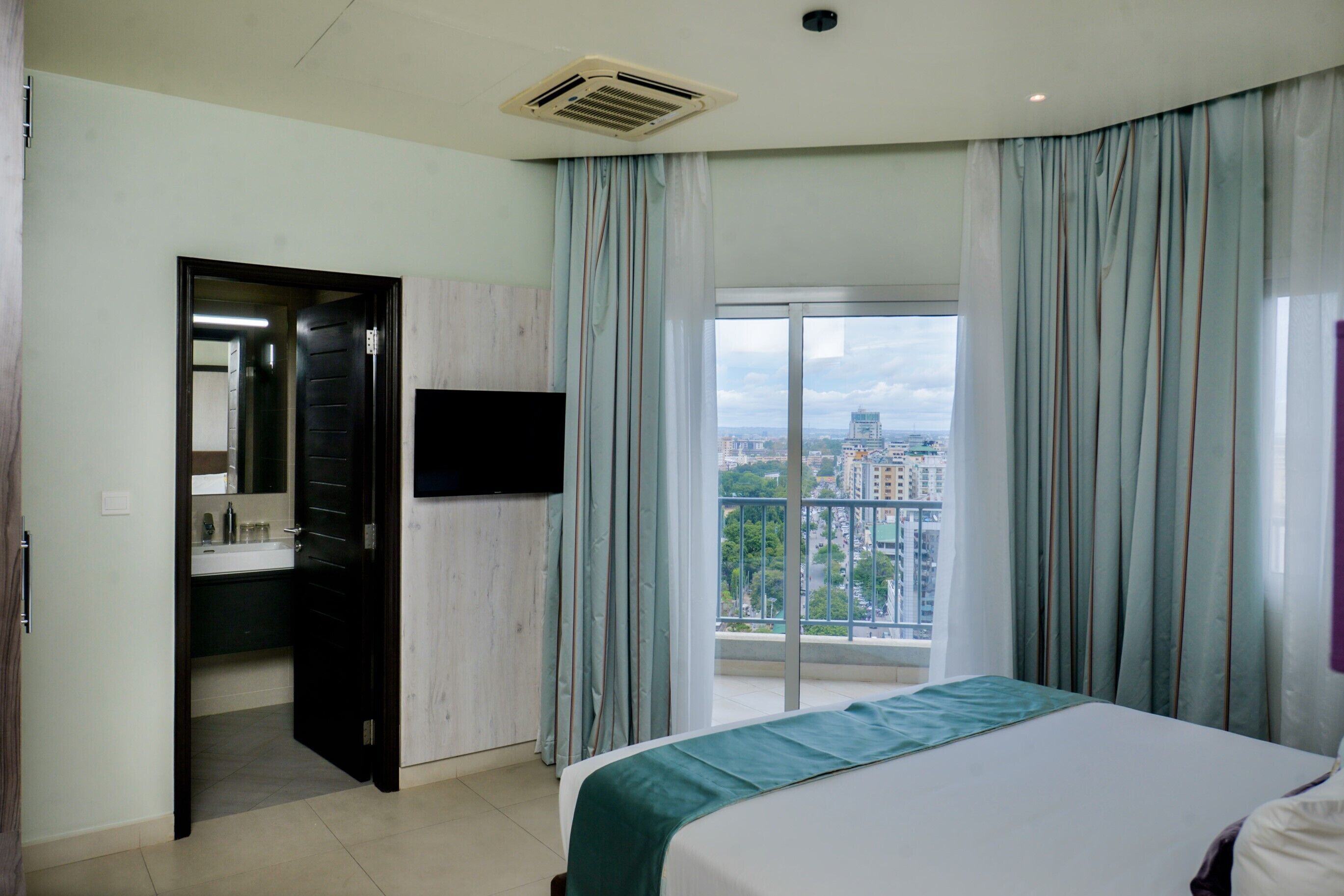 Habitación de hotel con cama, baño con espejo, lavabo y vista a un paisaje urbano a través de un ventanal grande.