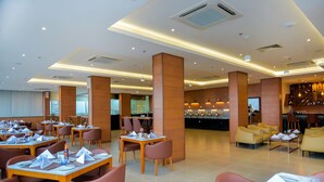 Un vaste espace de restauration dans l’hôtel, avec de nombreuses tables et chaises, un bar et, au fond, une vue sur la cuisine.