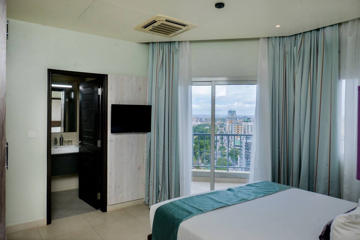 A hotel room with a bed, a bathroom with a mirror, a sink, and a view of a cityscape through a large window.