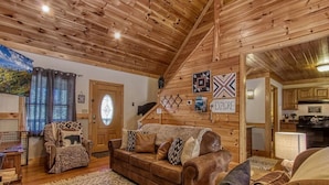 Un interior de cabaña acogedor con techo de madera, un sofá de cuero marrón, un tapete estampado y un comedor con mesa y sillas.