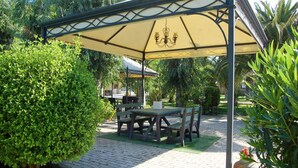 A covered patio area with a table and chairs, surrounded by greenery and a paved walkway.