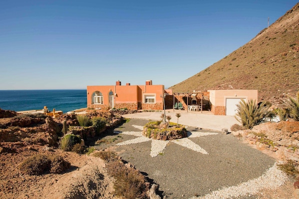 Une propriété située en bord de mer, avec une allée pavée en forme d’étoile menant à un bâtiment doté d’une terrasse et d’un espace salon extérieur.
