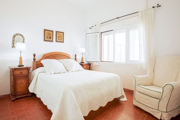 Um quarto com cama, mesas de cabeceira, uma cadeira e uma janela com cortinas.
