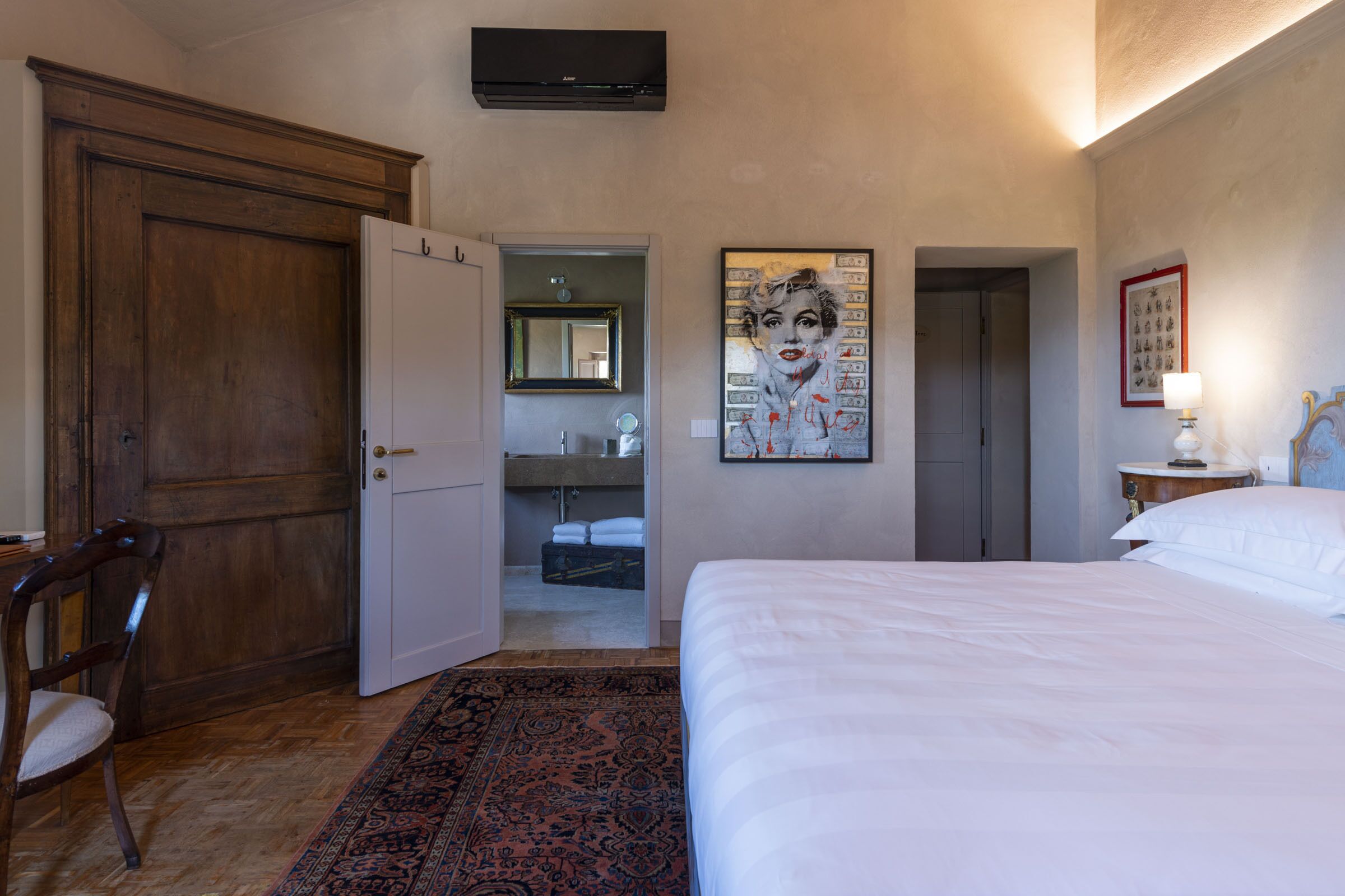 A hotel room with a large bed, a wooden door, a bathroom with a mirror, and a painting on the wall.