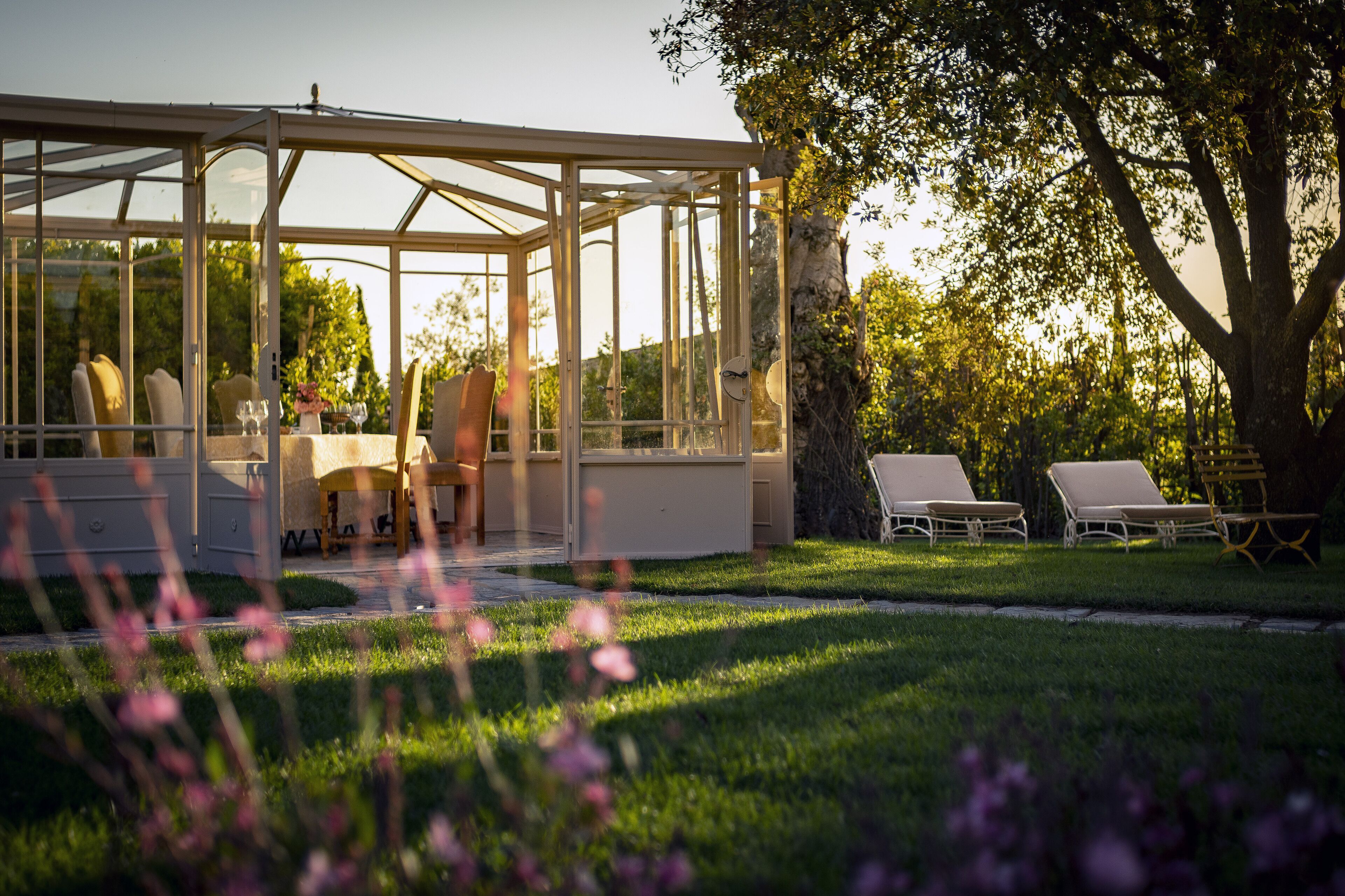 A conservatory with a glass roof, a dining table with chairs, and a garden with lawn chairs.