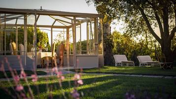 A conservatory with a glass roof, a dining table with chairs, and a garden with lawn chairs.