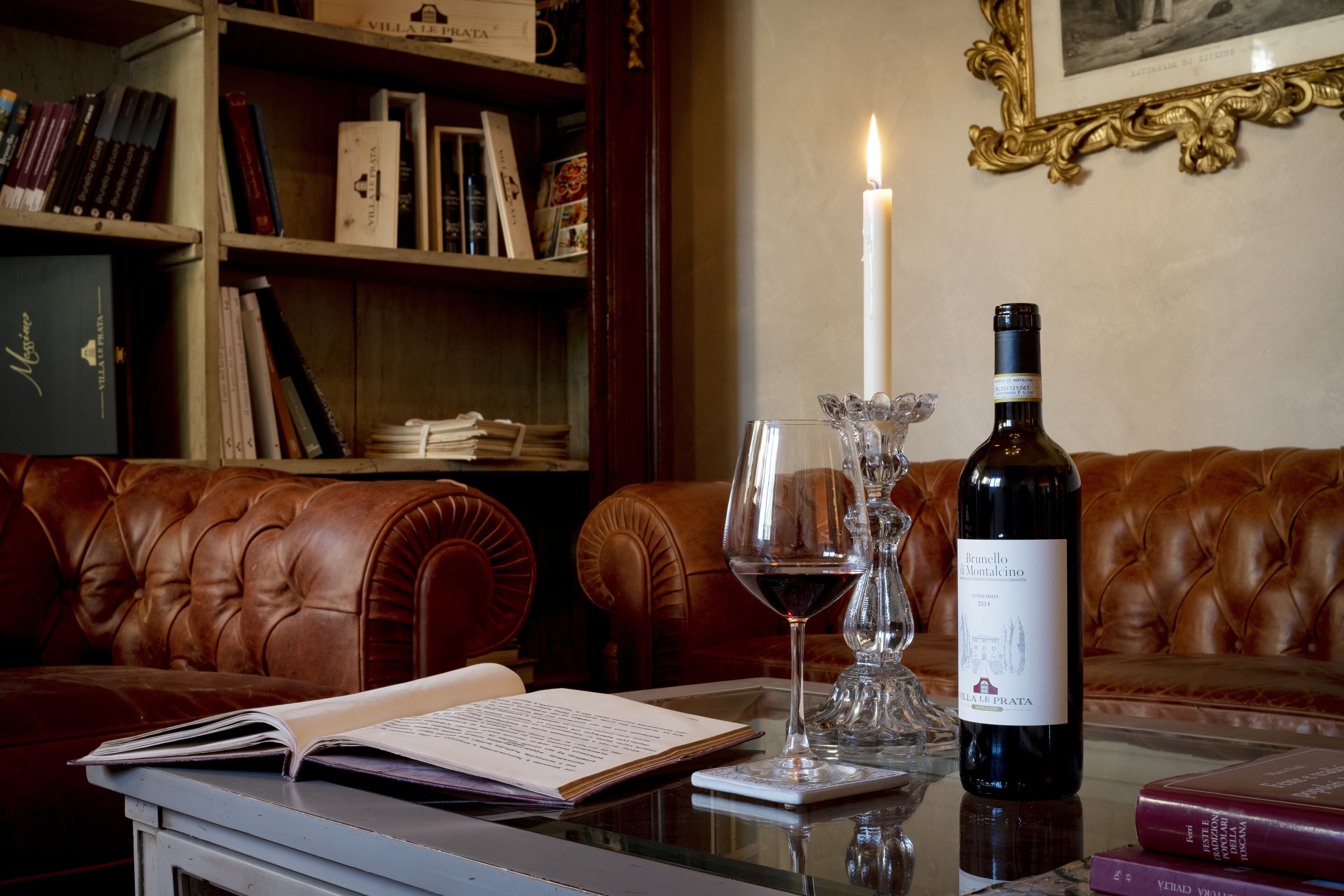 Un sofá de cuero, una mesa con una botella de vino y copas, y una estantería con libros y una vela.