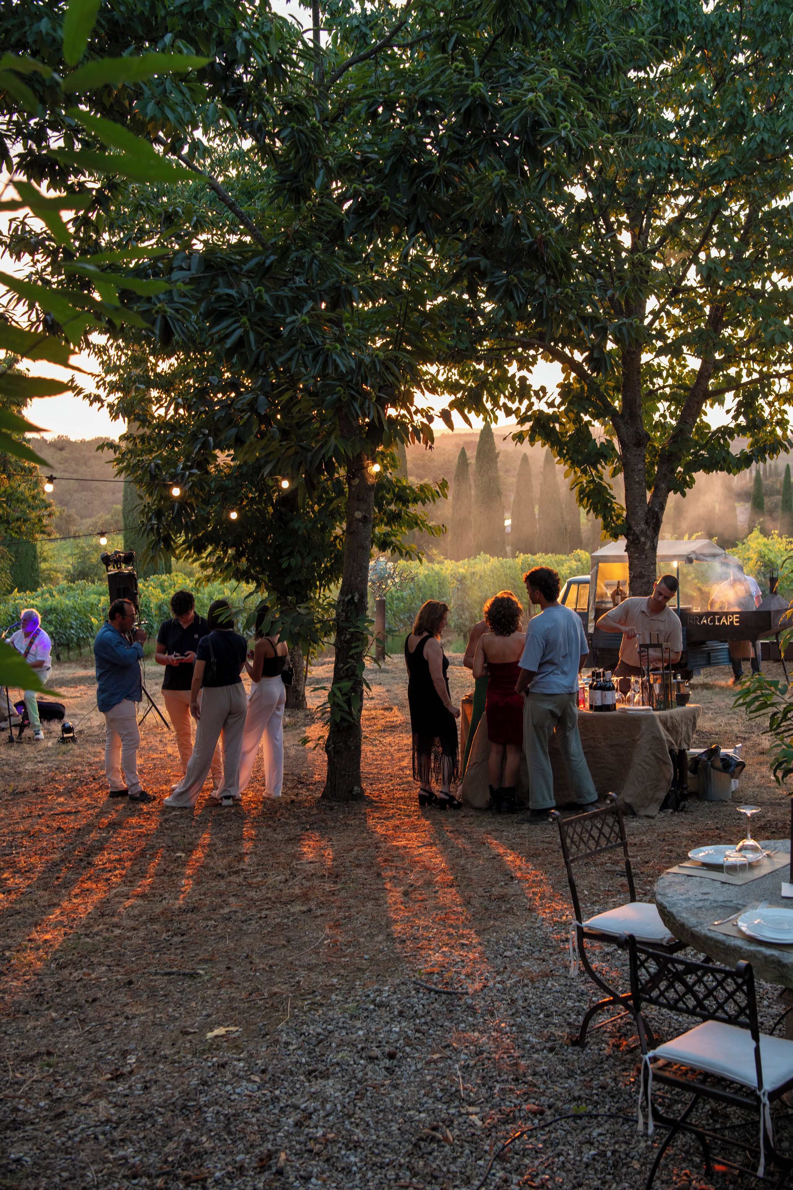 An outdoor gathering with people around a table under trees, a barbecue grill, and a tent in a garden setting.