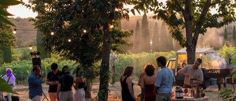 An outdoor gathering with people around a table under trees, a barbecue grill, and a tent in a garden setting.