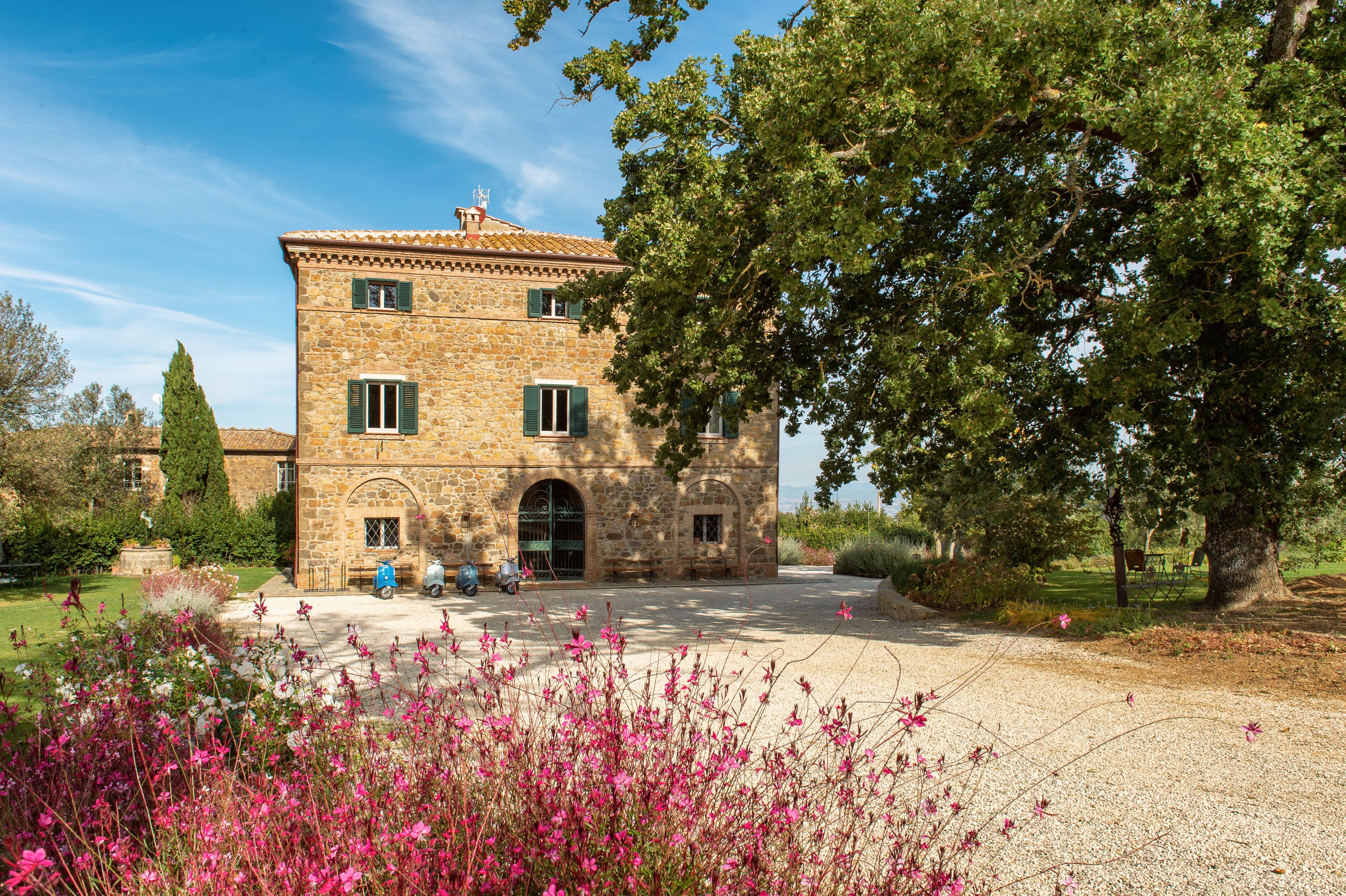 A stone building with a tiled roof, a central entrance, and a garden with pink flowers.