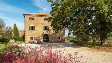 A stone building with a tiled roof, a central entrance, and a garden with pink flowers.