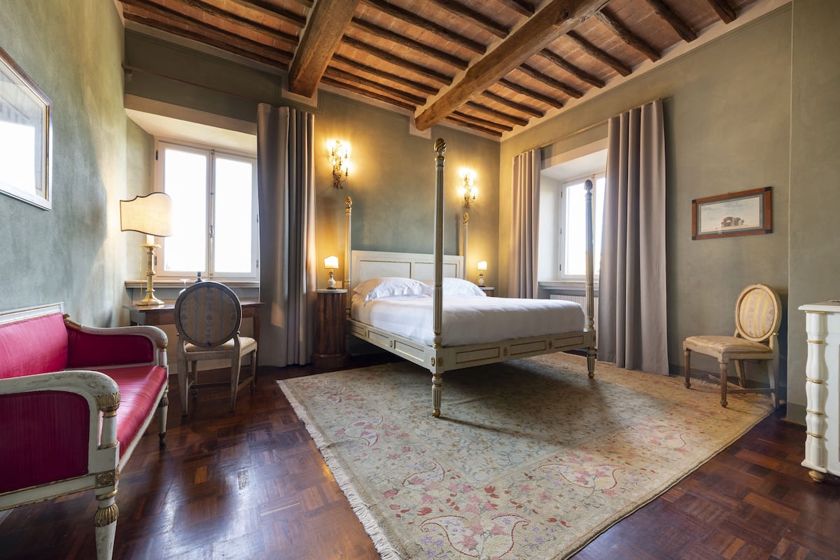 A bedroom with a four-poster bed, a red sofa, two chairs, a large window, and a wooden ceiling.