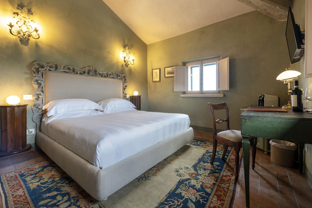 A bedroom with a large bed, a desk, and a chair. There is a window with white shutters and a lamp on the desk.